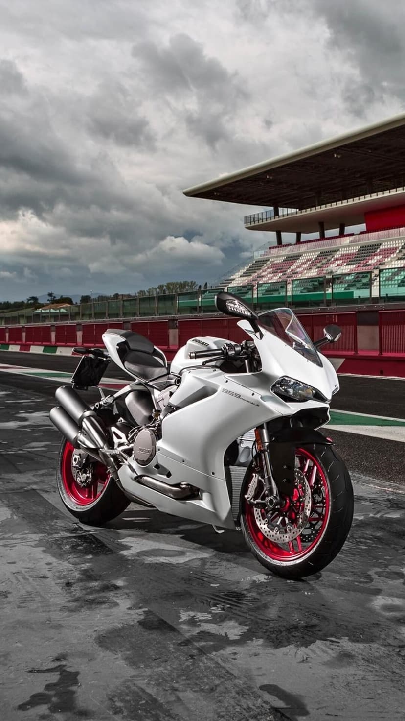 White Ducati Panigale motorcycle with red wheels parked on a wet racing track beneath a cloudy sky and grandstands.