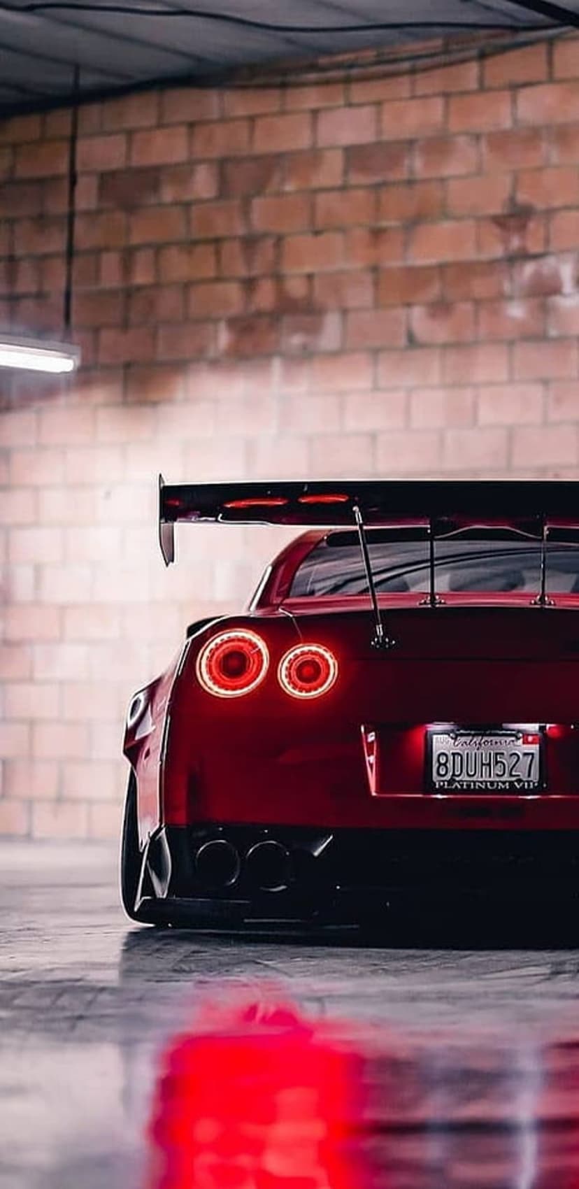 Red Nissan GT-R rear view with a black wing and circular taillights parked on a reflective garage floor.