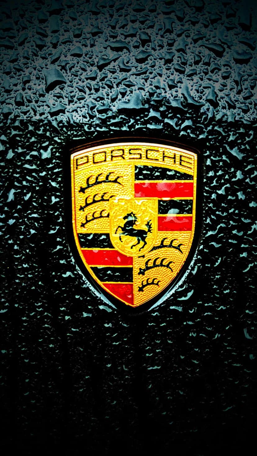 Porsche crest with gold and red enamel sits on a dark textured surface covered in beads of water droplets.