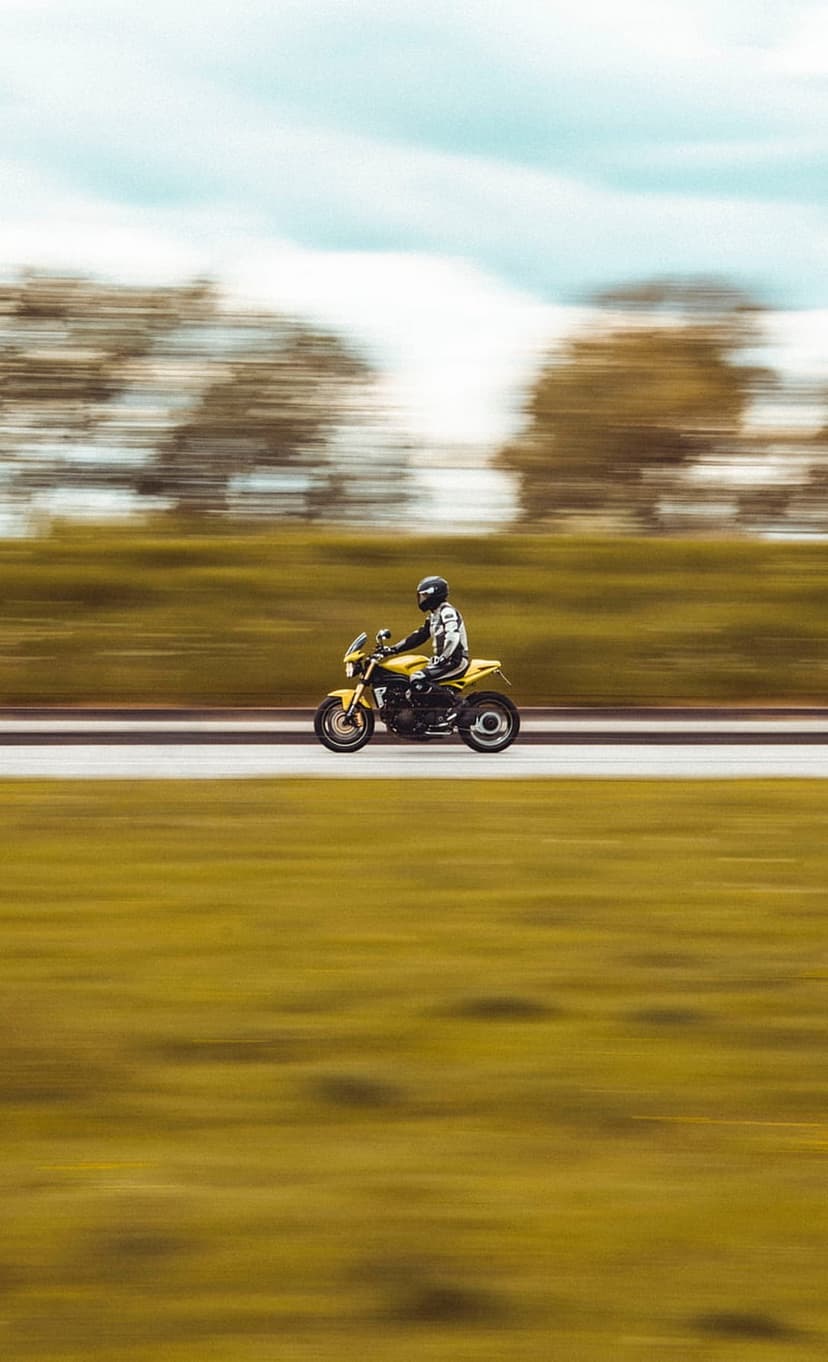 Motorcyclist wearing yellow gear riding fast on a road with motion blurred green trees and a soft blue sky.