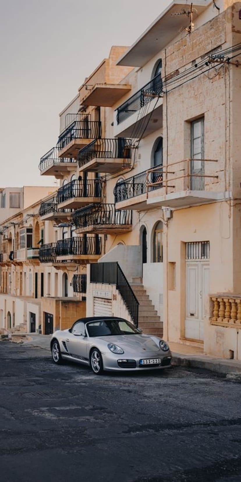 Silver Porsche Boxster convertible on a cobblestone street with Mediterranean balconies and arched windows.