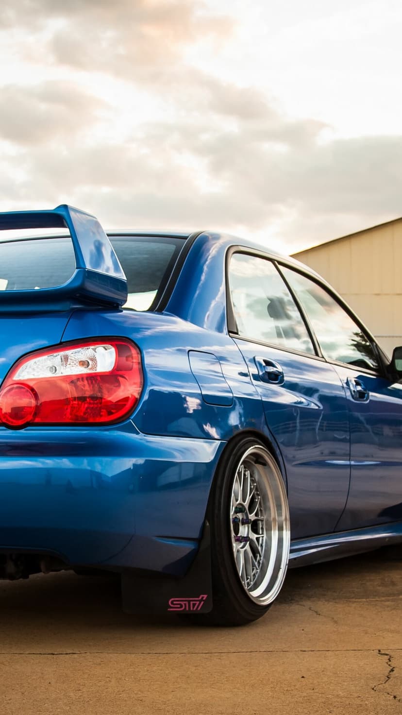 Rear view of a blue Subaru WRX STI with custom silver wheels and a pink logo on the mudflap at sunset.
