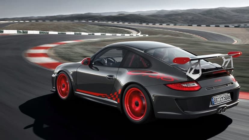 Dark grey Porsche 911 GT3 RS with red wheels and decals racing on a winding asphalt track with motion blur.