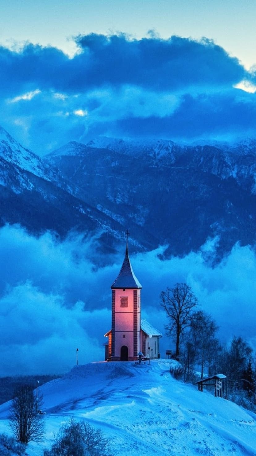 White steeple church on a snowy hill with misty blue alpine mountains and a pink dawn sky in the background.