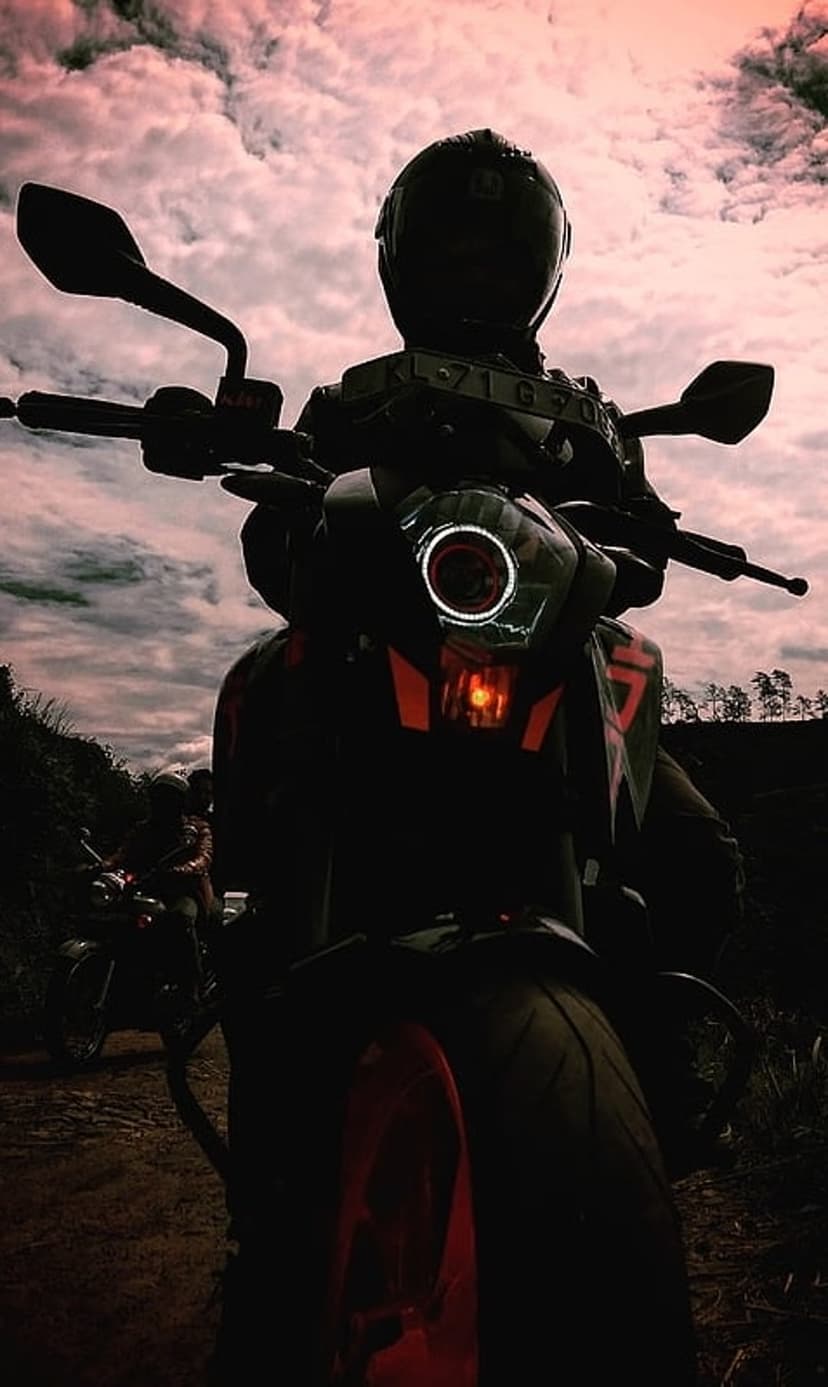 Lone motorcyclist silhouette with a glowing orange headlight against a pink and grey sunset cloudscape.