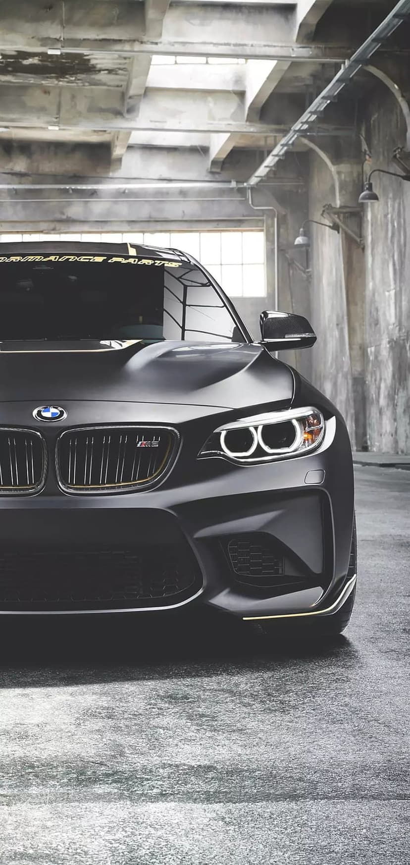 Matte black BMW M2 front view with LED headlights in a dark industrial garage and wet asphalt reflections.
