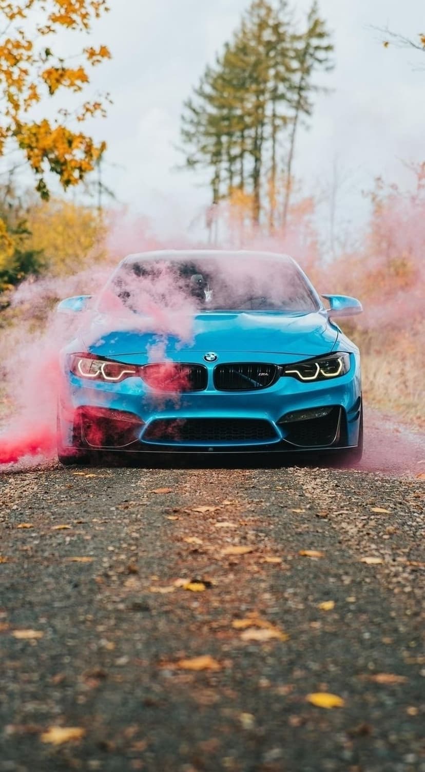 Blue BMW M4 sports car emitting bright pink tire smoke on a gravel road surrounded by orange autumn trees.