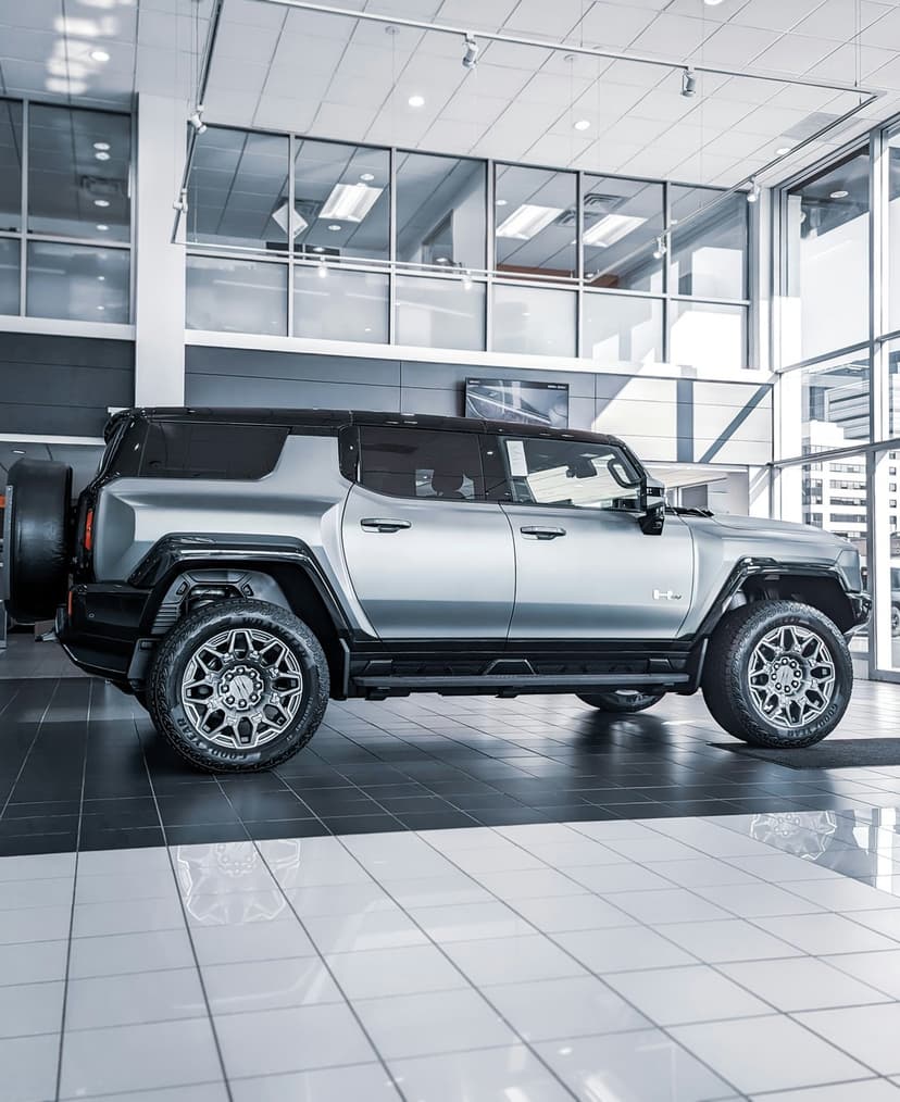 Silver GMC Hummer EV SUV parked on a reflective showroom floor near large windows with bright natural lighting.