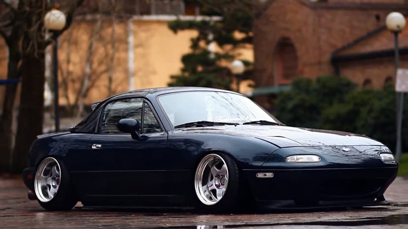 Dark blue Mazda Miata NA with extreme camber and silver wheels parked on a wet, reflective brick road.