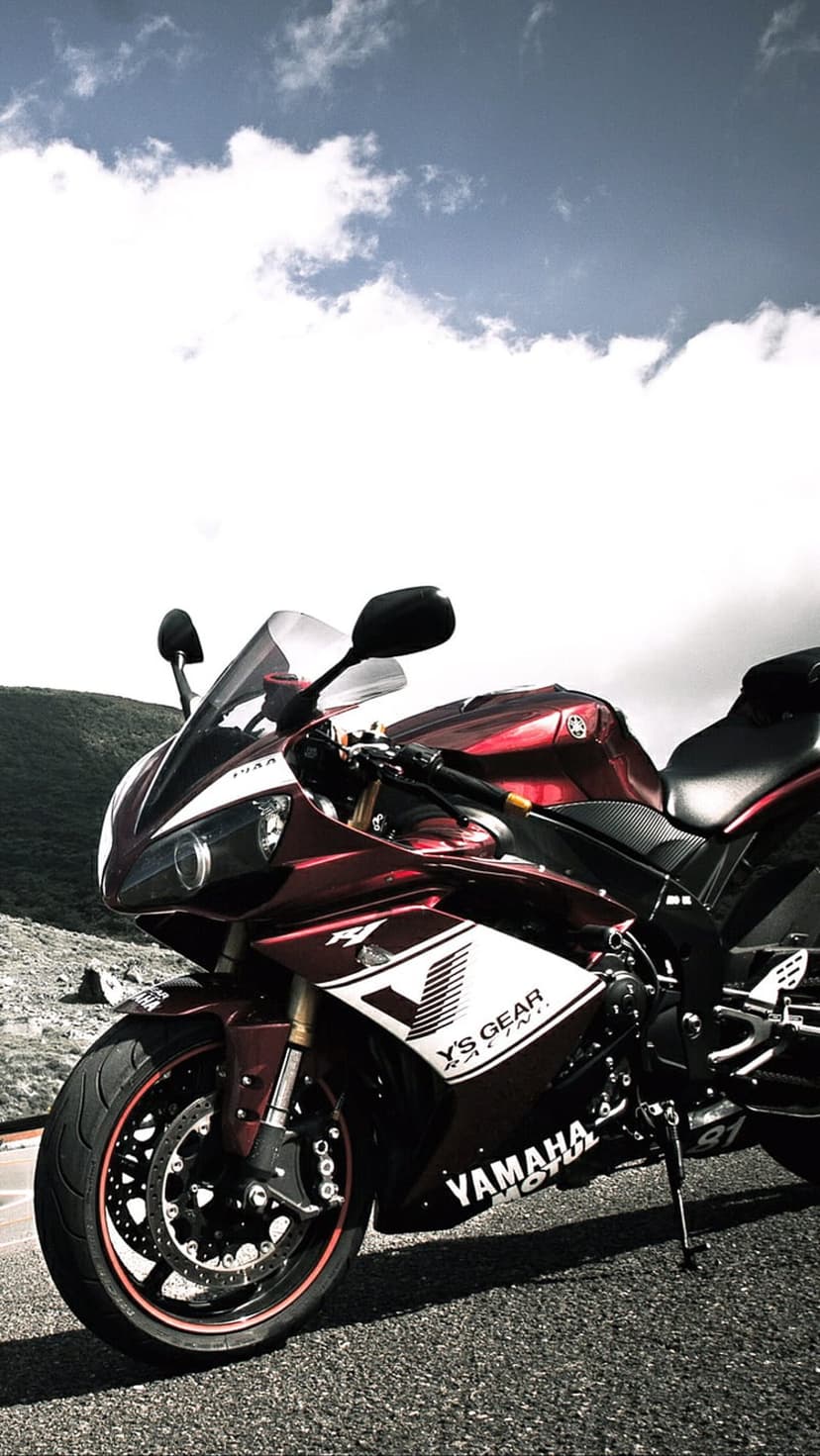 Red Yamaha R1 sportbike parked on a mountain road under a cloudy sky with green hills in the background.