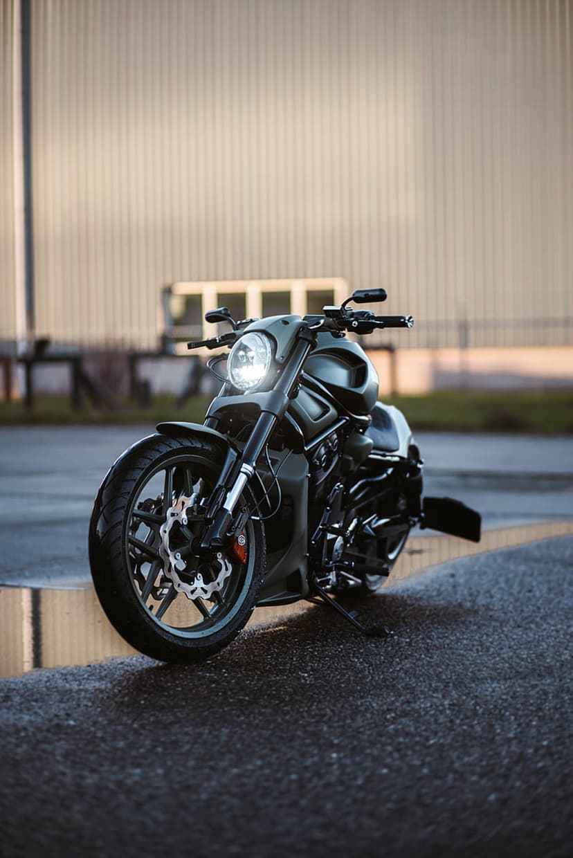 Black Harley Davidson motorcycle parked on wet asphalt with a glowing headlight and industrial background.