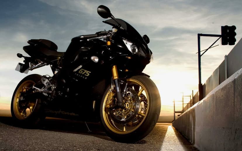 Black Triumph Daytona 675 motorcycle with gold wheels parked on a track under a golden sunset sky.