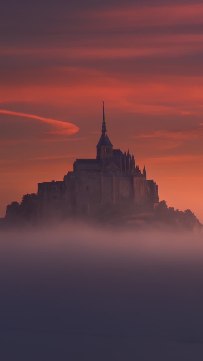 Mont Saint-Michel abbey silhouette rises above thick white mist under a vibrant orange and red sunset sky.