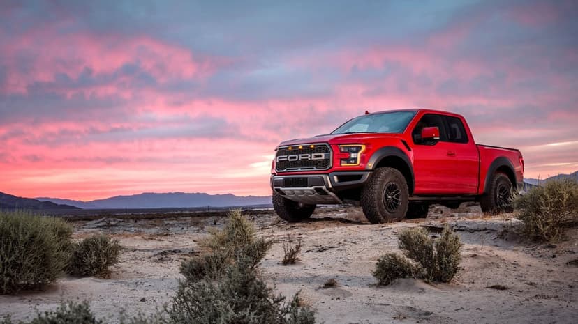 Red Ford Raptor truck on a sandy desert hill under a vibrant pink and purple sunset sky with distant mountains.