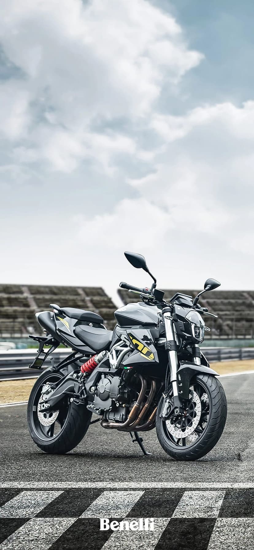 Black Benelli motorcycle on a racetrack with a red shock absorber, blurred stadium, and cloudy sky overhead.