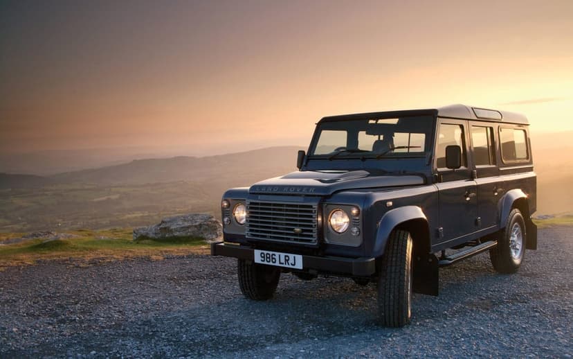 Blue Land Rover Defender parked on a gravel hilltop overlooking misty green mountains during a golden sunset.