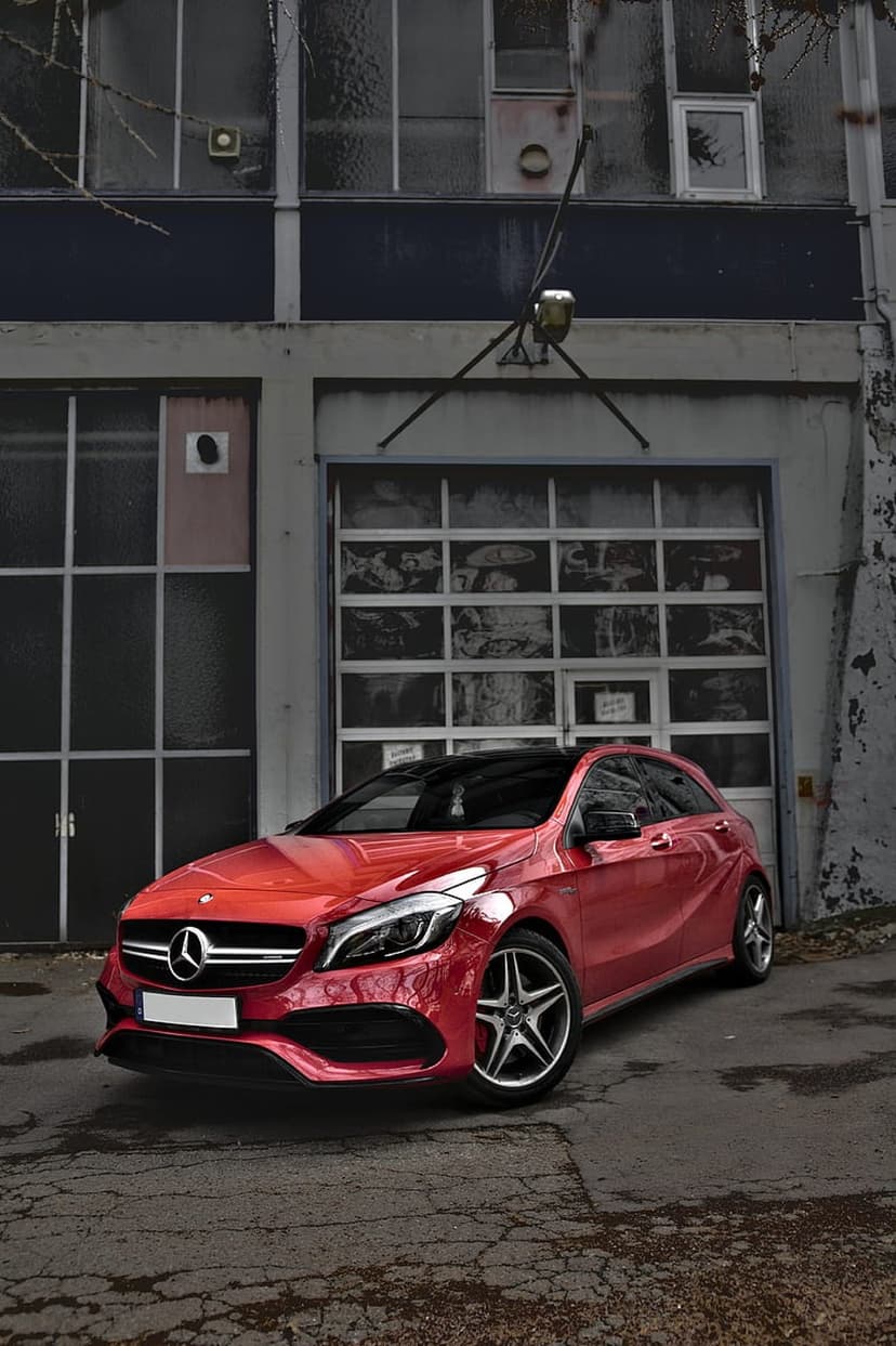 Crimson Mercedes-Benz A-Class hatchback parked before a large industrial garage door at an urban site.