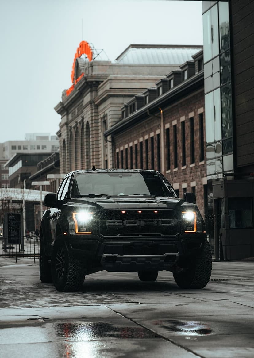 Black Ford F-150 Raptor truck with glowing headlights parked on a wet street reflecting city buildings.