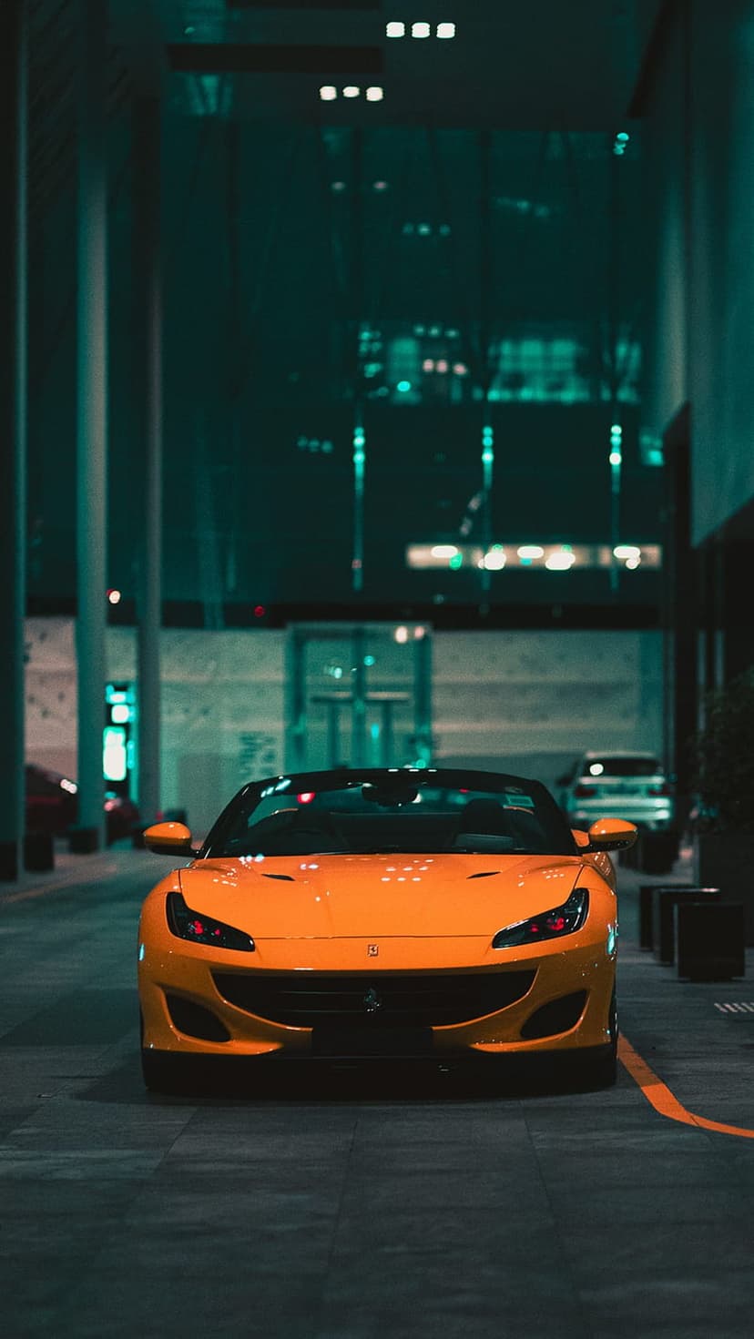 Orange Ferrari convertible on wet asphalt at night under glowing city lights in a low-angle vertical shot.