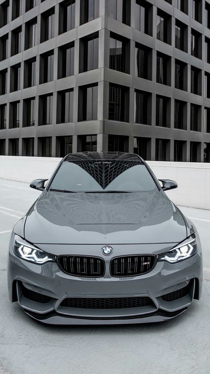 Grey BMW M4 sports car parked in front of a modern glass building with sharp architectural lines and reflections.