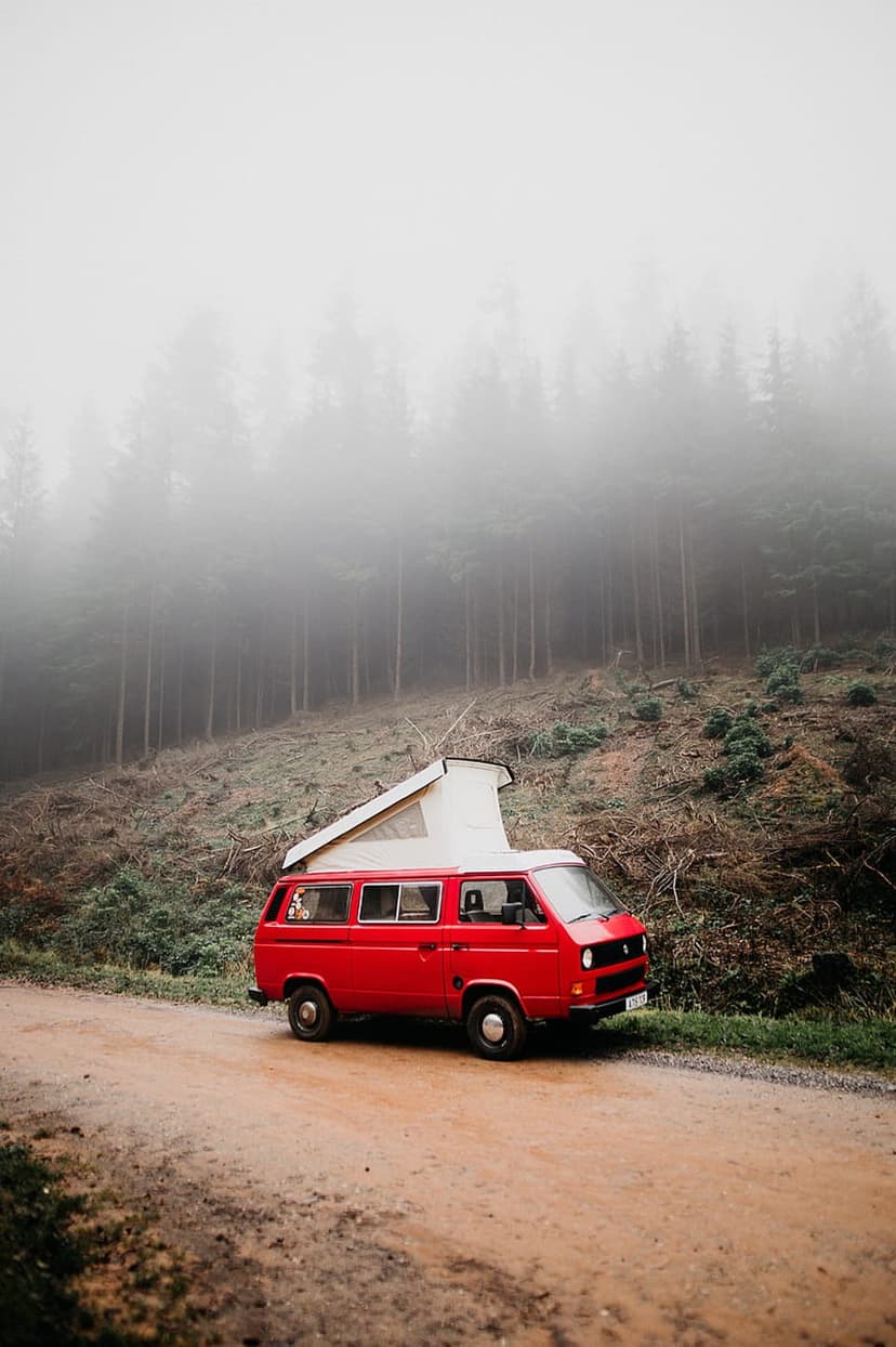 Vintage red camper van with a pop-top roof parked on a muddy forest road under thick, atmospheric fog.