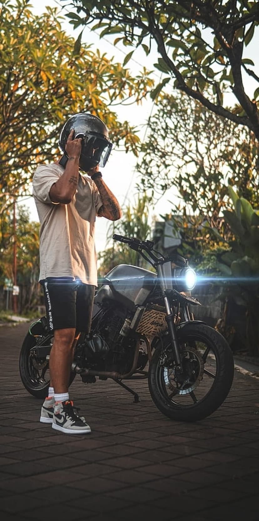 Man putting on a black helmet beside a motorcycle with an illuminated headlight in a sunlit outdoor setting.