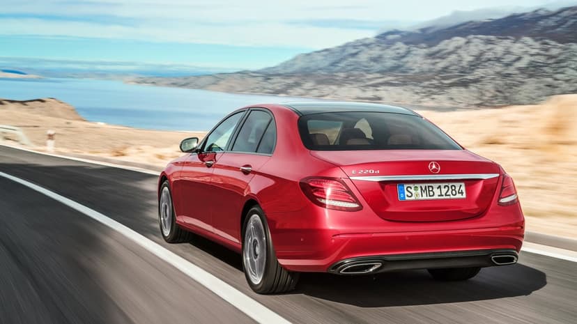 Red Mercedes-Benz E-Class sedan driving on a coastal road with blue water and mountains behind.