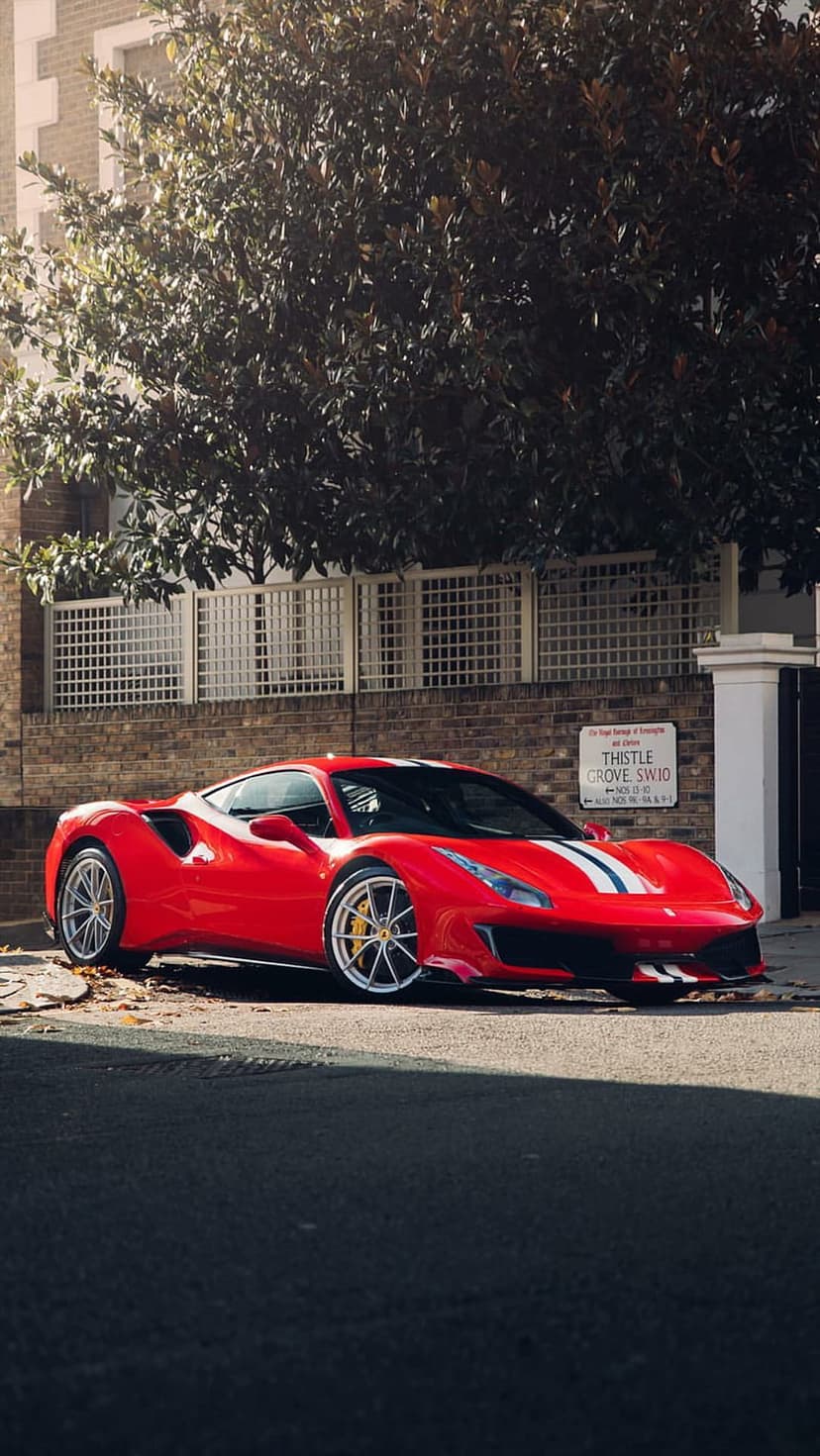 Red Ferrari sports car with racing stripes parked on an asphalt street under the shade of a large green tree.