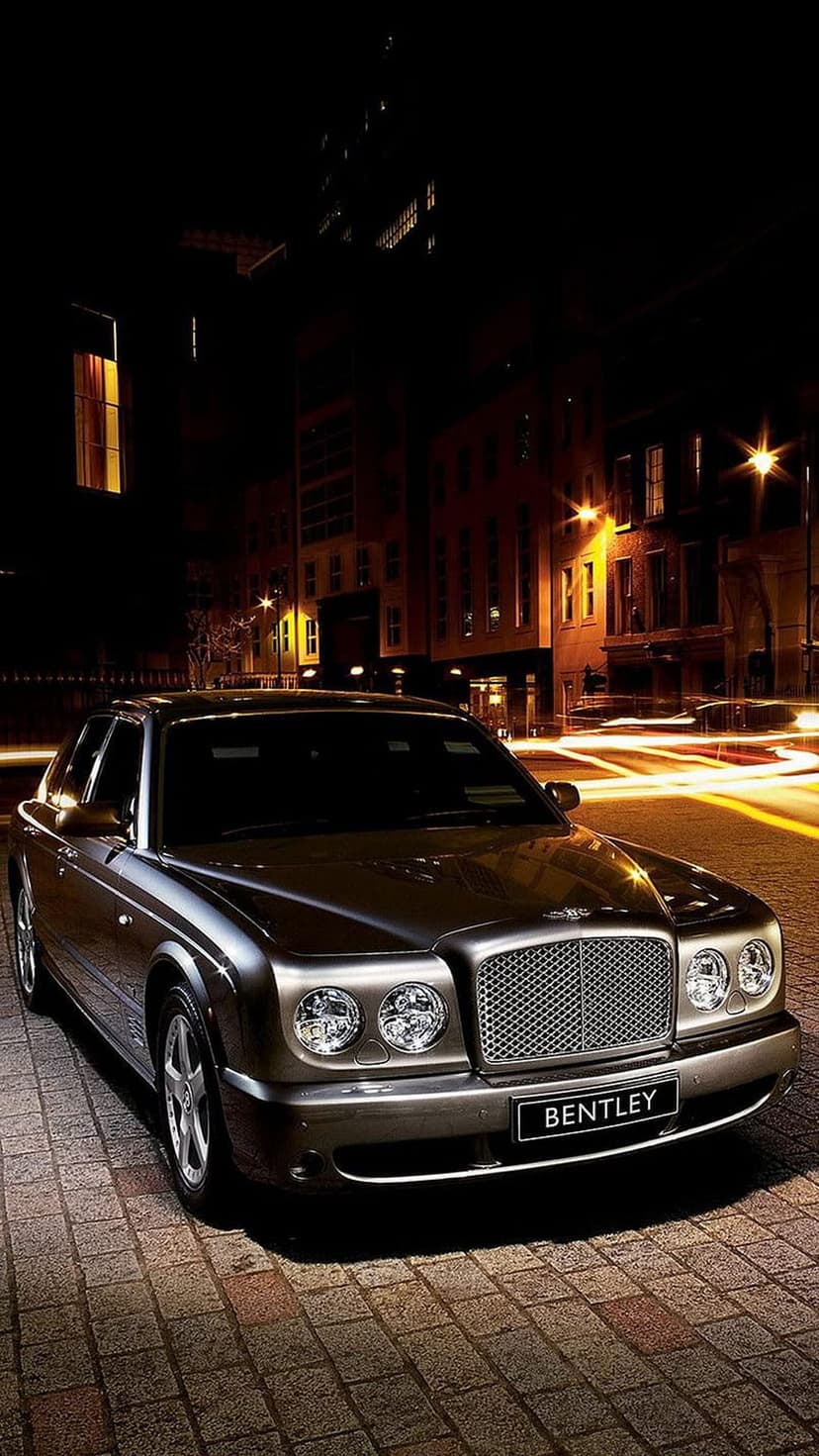 Silver Bentley sedan with glowing headlights parked on wet cobblestone streets under warm city lights at night.