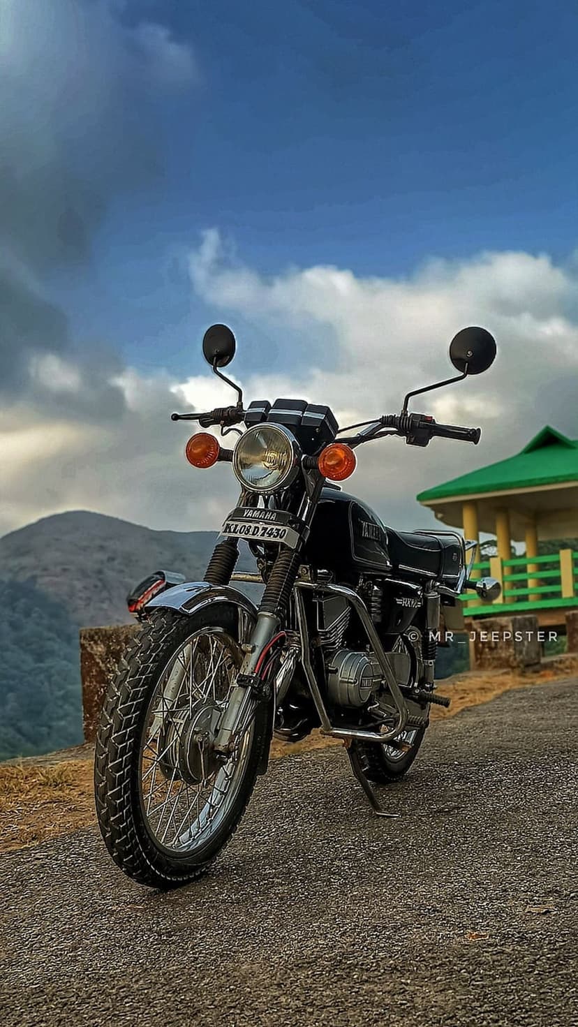 Black Yamaha RX 100 vintage motorcycle on a gravel road with misty green mountains under a cloudy gray sky.
