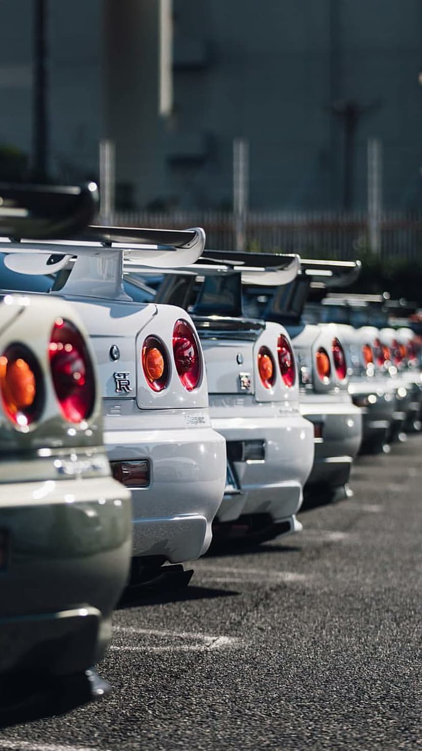 Nissan Skyline R34 GT-R taillights in a row on white cars parked on sunlit asphalt in a vertical perspective.