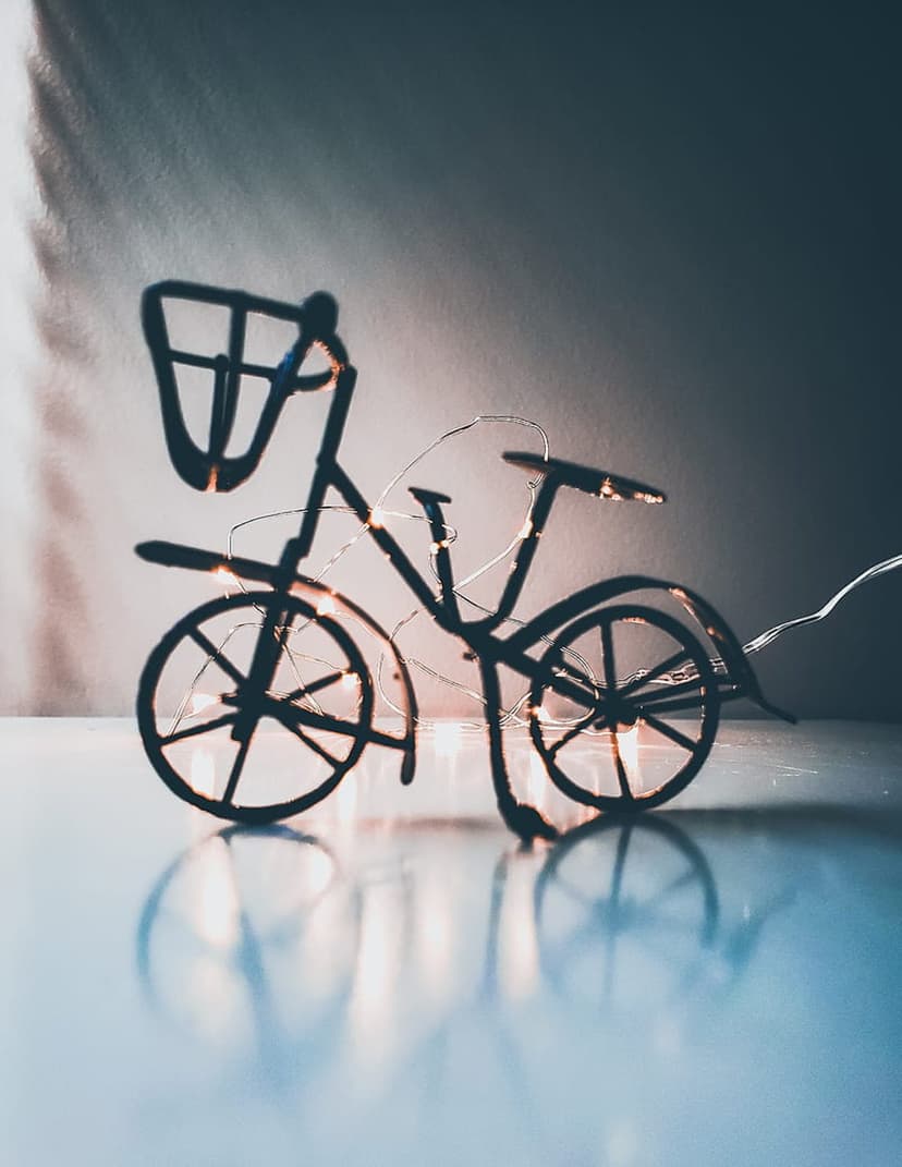 Wire bicycle miniature with golden fairy lights reflecting on a glossy dark surface in a vertical composition.