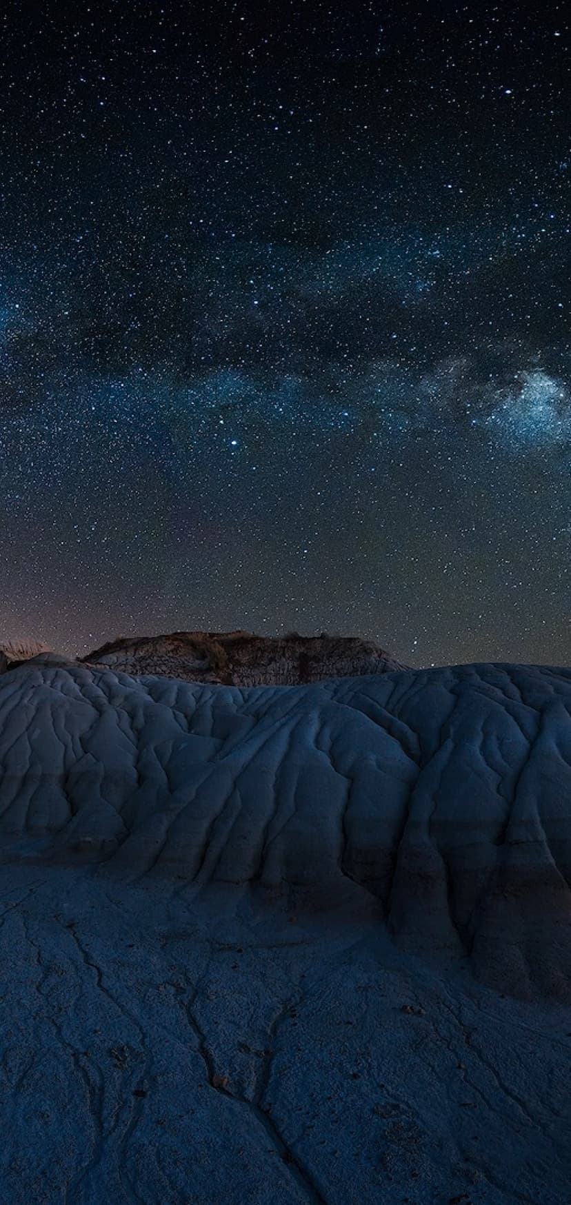 Milky Way galaxy stretching over blue-grey sedimentary rocks and dark mountain ridges under a starry night sky.