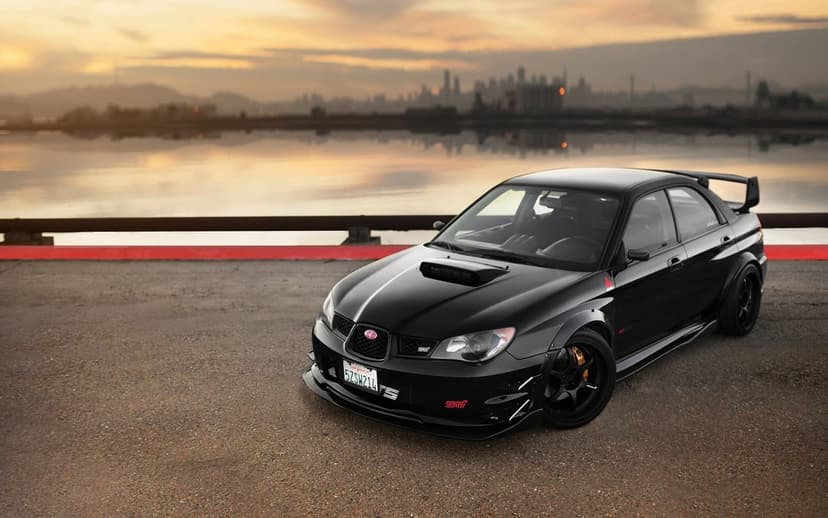 Black Subaru Impreza WRX STI with a widebody kit parked near a bay at sunset with a distant city skyline.