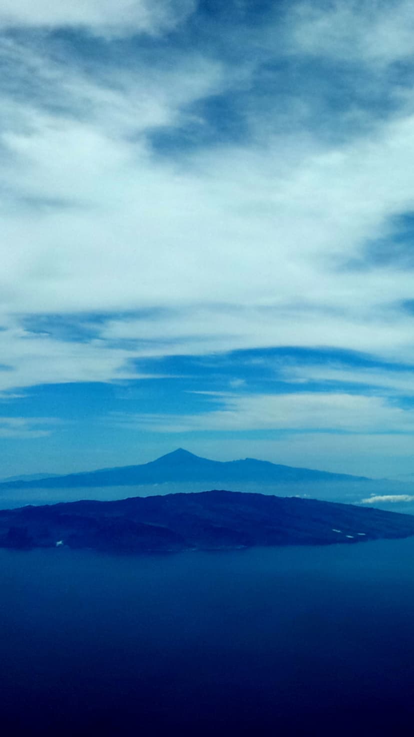 Dark island silhouettes and distant mountains rest on a vast blue lake surface under a pale, hazy sky.