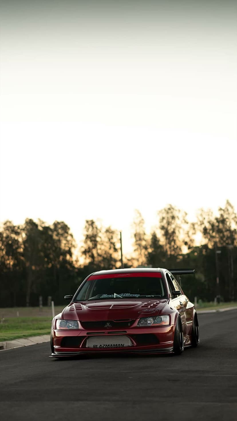Red Mitsubishi Lancer Evolution X with a large rear wing and front intercooler sits on an asphalt forest road.