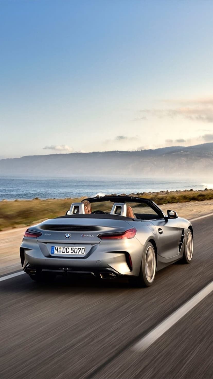 BMW Z4 silver convertible driving on a winding coastal road beside blue ocean waves and lush green hills.