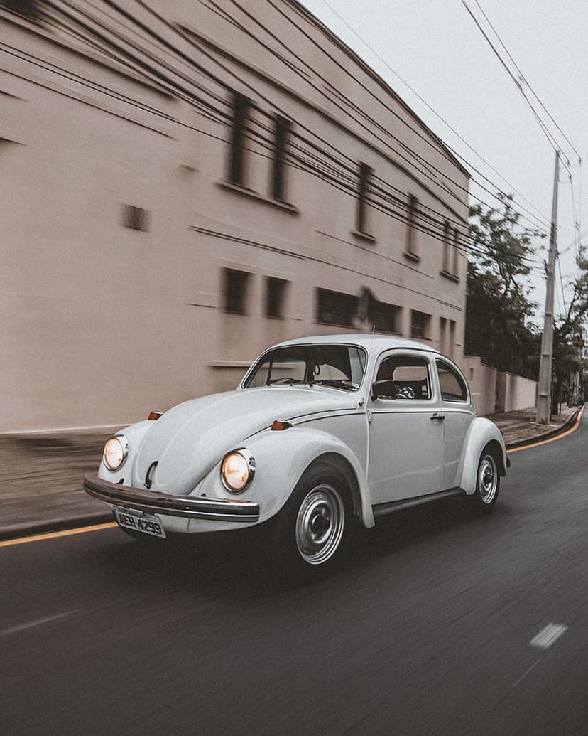 White Volkswagen Beetle with lit headlights drives down a tree-lined street with motion blur at sunset.
