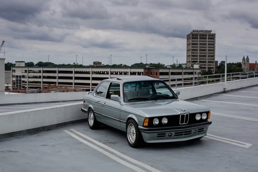 Silver BMW E30 coupe parked in a lot with a concrete barrier and a multi-story garage under an overcast sky.