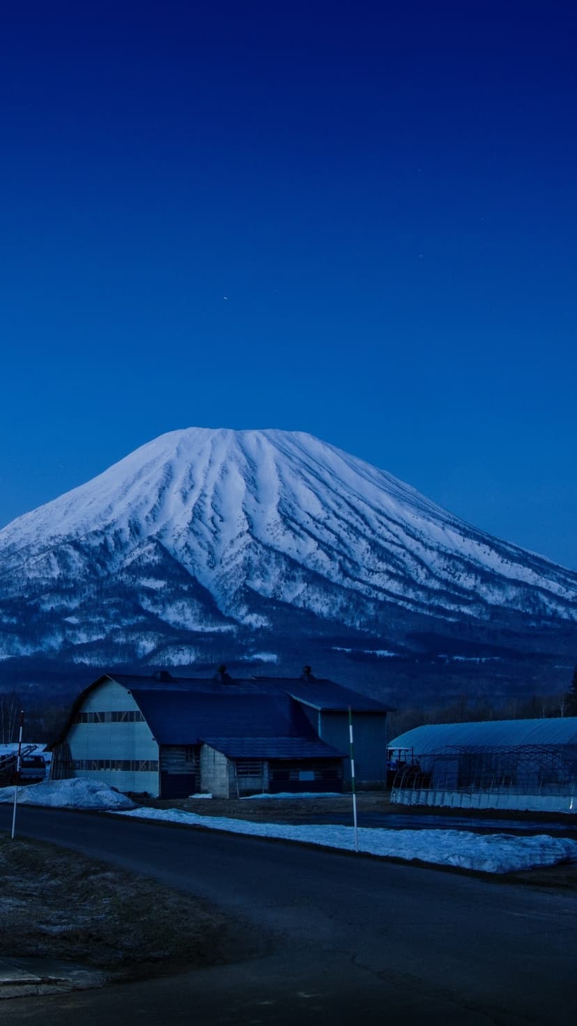 Snow-covered mountain peak rising behind small farm buildings under a clear blue twilight sky in rural Japan.