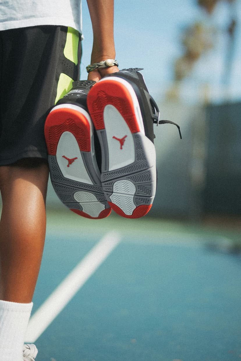 Person holding red and black Jordan sneakers over a blue tennis court, revealing the Jumpman logo on the soles.