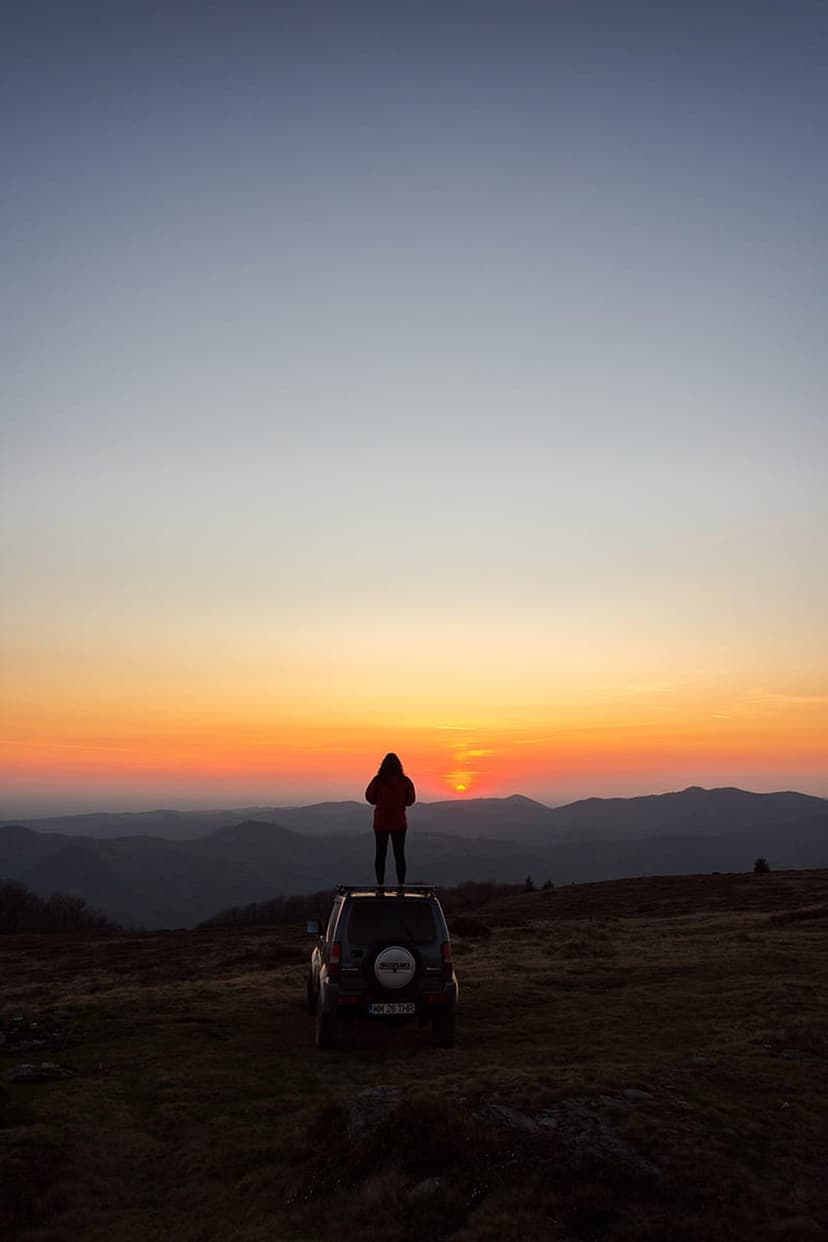 Person silhouetted on a car roof gazing at distant mountains under a vibrant orange and blue sunset sky.