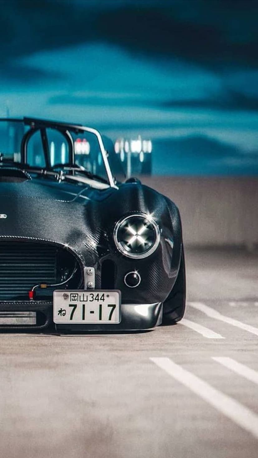 AC Cobra sports car with carbon fiber body and Japanese license plate parked on a night street under blue light.