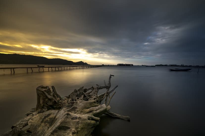 Weathered driftwood sits near a long wooden pier extending into calm water under a dramatic sunset and storm clouds.
