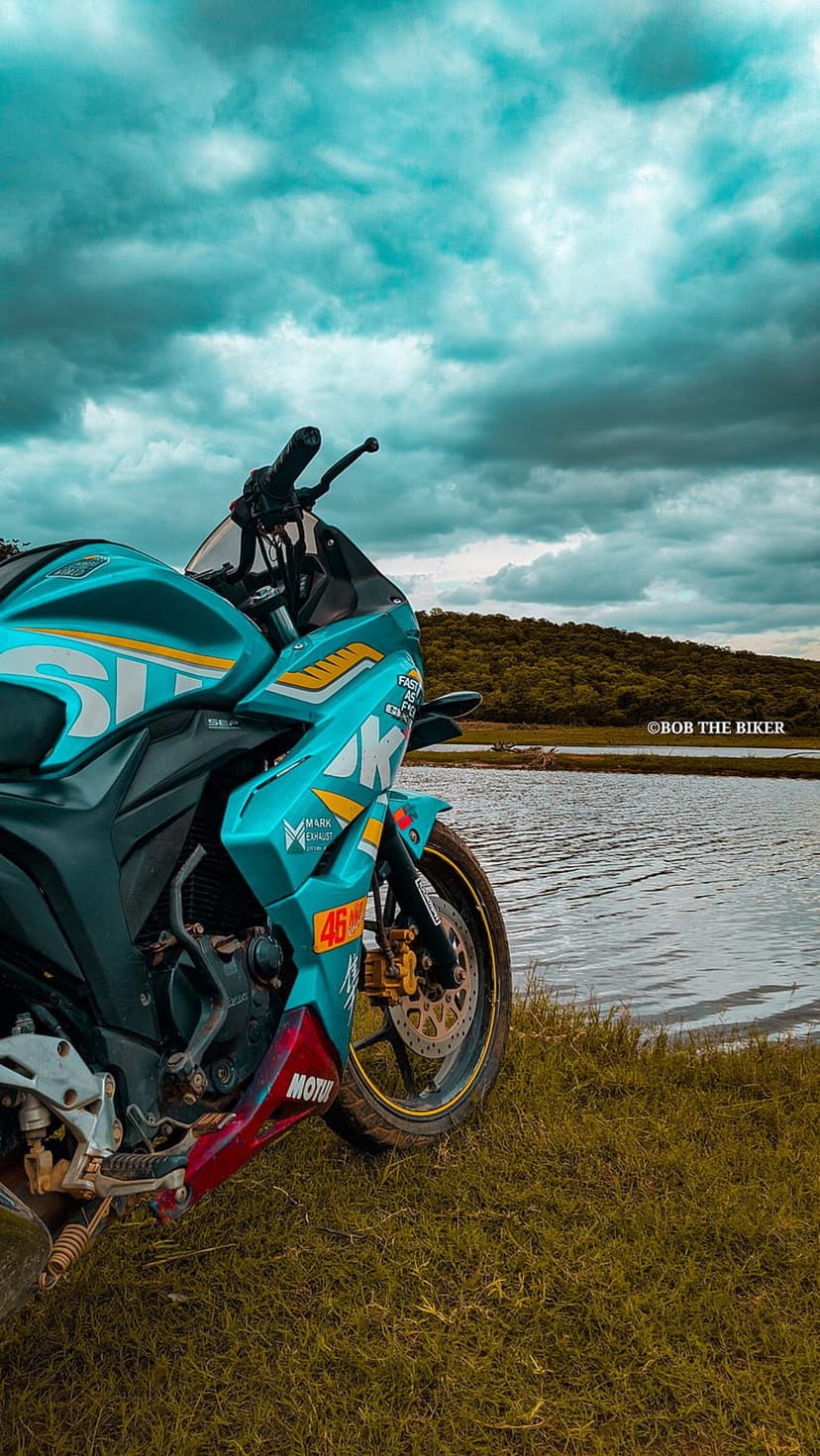 Turquoise and red sport motorcycle parked on green grass beside a calm lake under a dark, cloudy sky.