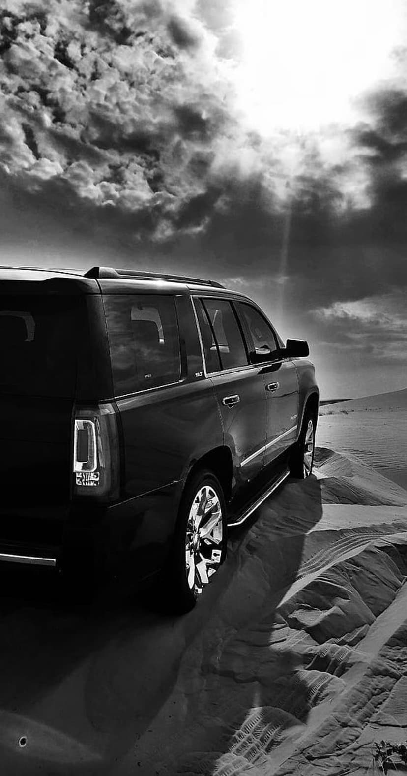 Black SUV parked on rippling sand dunes under a cloudy sky with sunbeams in a high-contrast monochrome style.