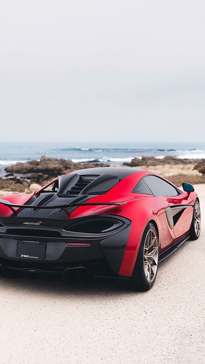 Red McLaren supercar on an asphalt road next to a rocky shoreline under a cloudy sky with ocean waves.