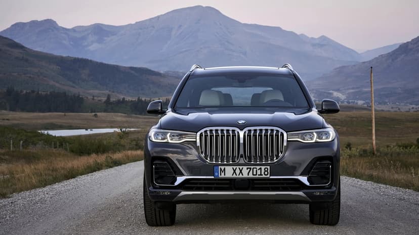 BMW X7 luxury SUV parked on a gravel road with a scenic mountain range and lake in the background at dusk.