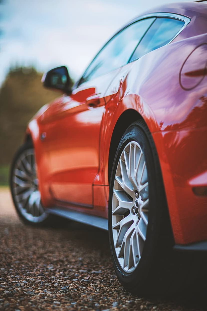 Red Ford Mustang rear quarter panel and silver alloy wheel resting on a textured gravel surface in natural light.