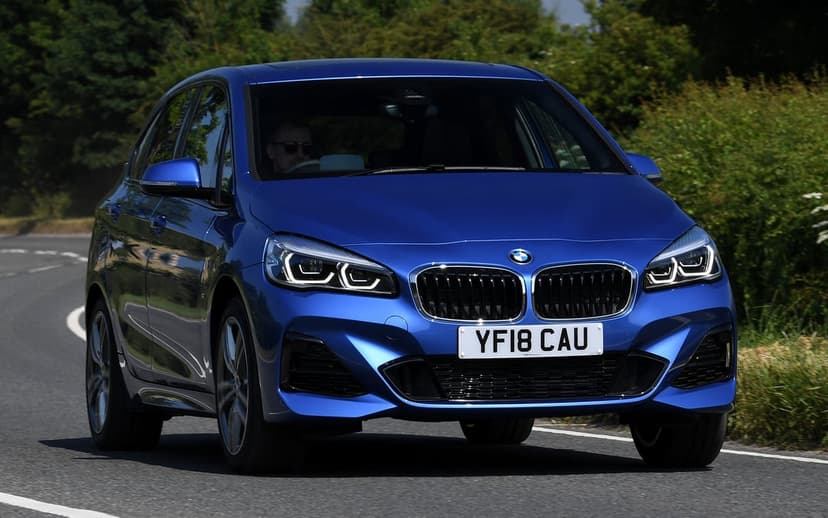 Blue BMW 2 Series car driving on a paved road surrounded by green trees under bright daytime sunlight.